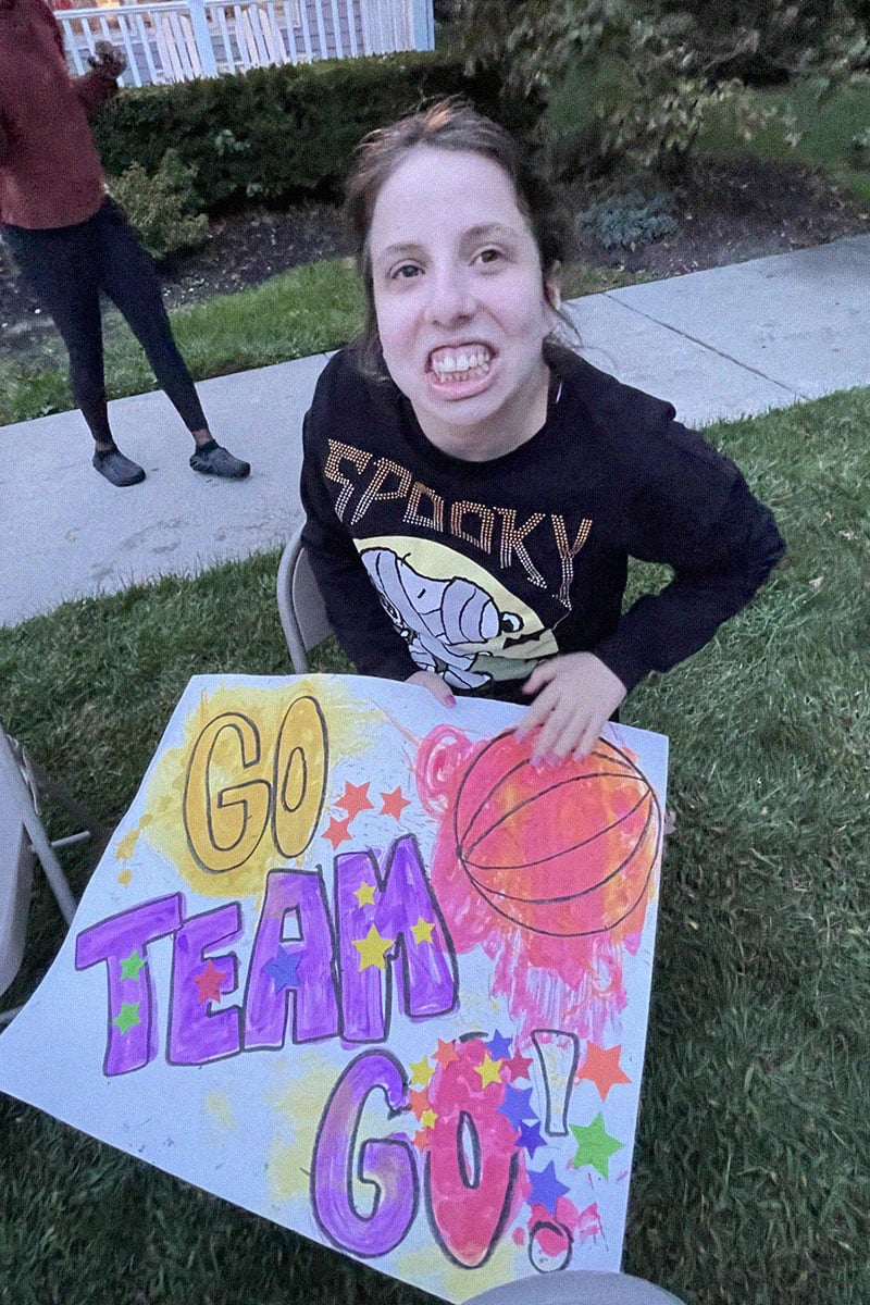 female sitting in chair on grass holding sign that says go team go