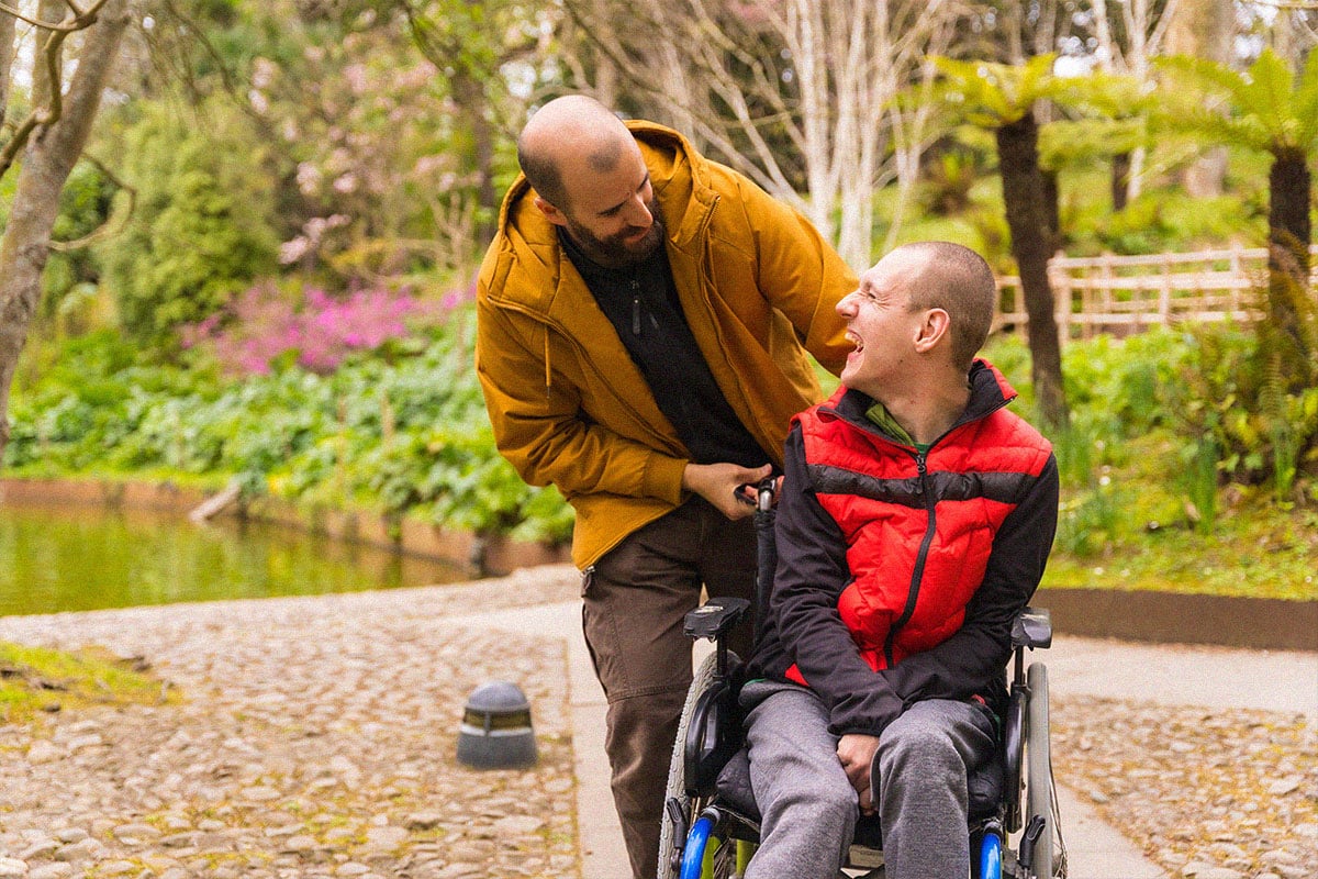 person with a disability in the wheelchair being pushed by a friend in a public city park